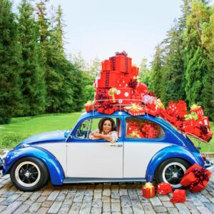 Oprah Winfrey sits in a VW Beetle covered in red presents with evergreens in the background