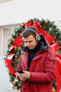 White male-presenting individual with short brown hair wearing red winter jacket and gold ring is holding and looking down at cell phone and appears to be listening to music through attached headphones, large green wreath with red bow in background