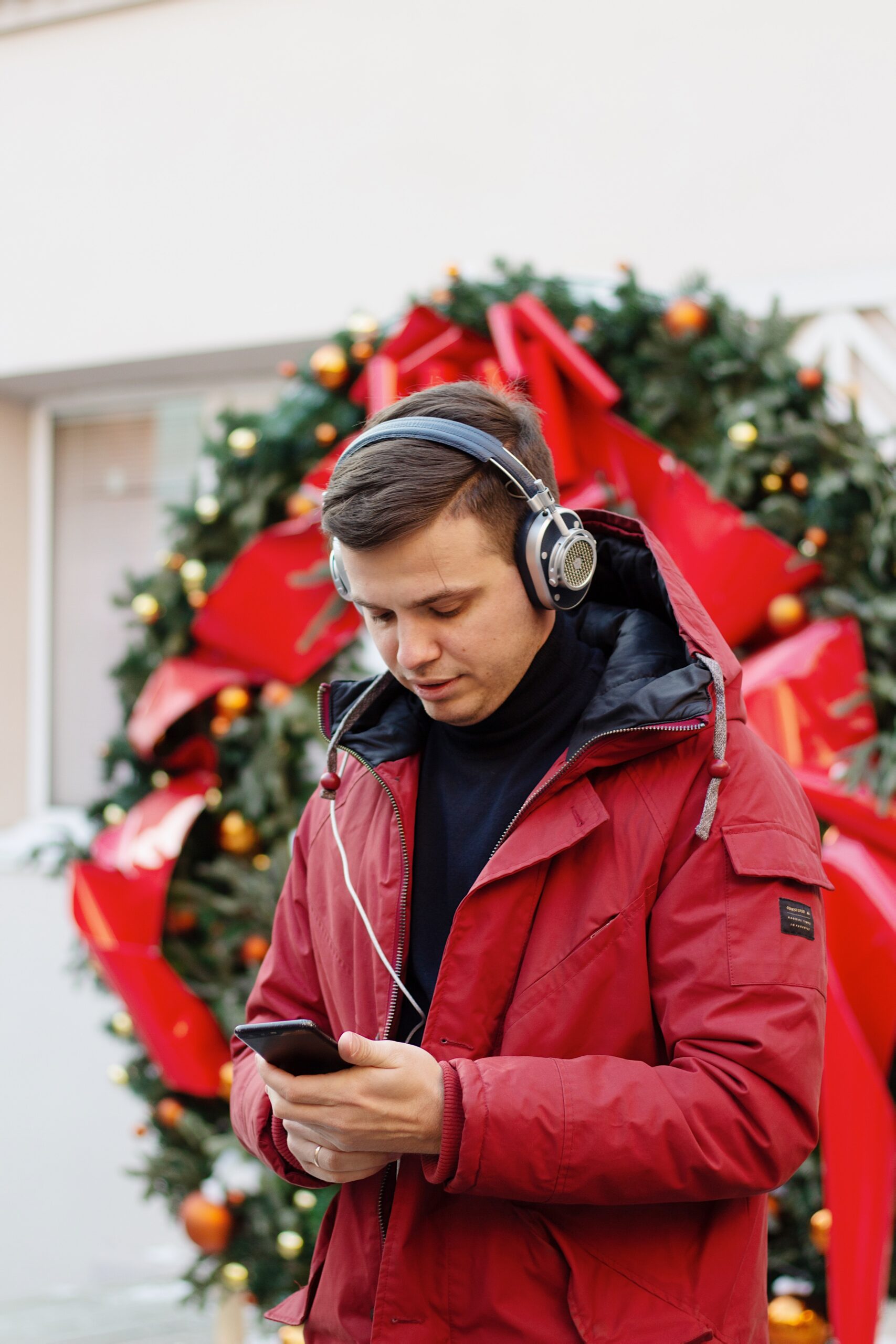 White male-presenting individual with short brown hair wearing red winter jacket and gold ring is holding and looking down at cell phone and appears to be listening to music through attached headphones, large green wreath with red bow in background